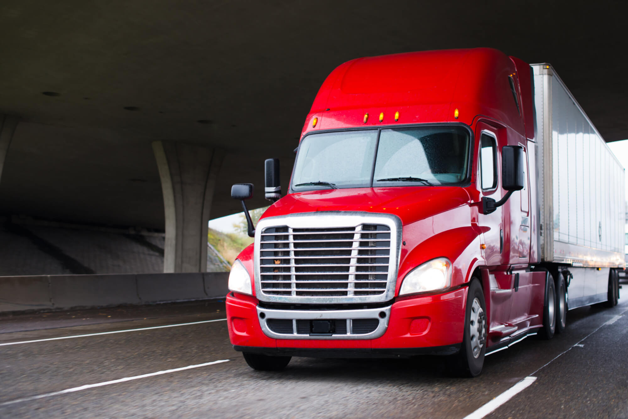 A modern big rig semi truck for long haulage with a high cabin for improving aerodynamic characteristics moves under the bridge across a multi-lane highway transporting a dry van semi trailer with commercial cargo to the place of delivery