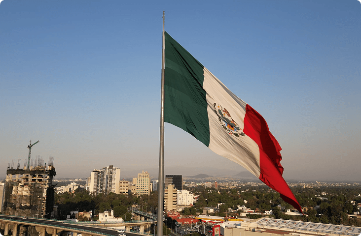 A Mexico flag blowing in the breeze with an industrial city and blue sky in the background.
