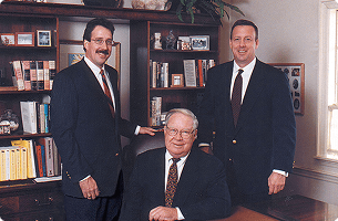 A picture of two people standing behind one person sitting at a formal desk with a bookcase in the background.