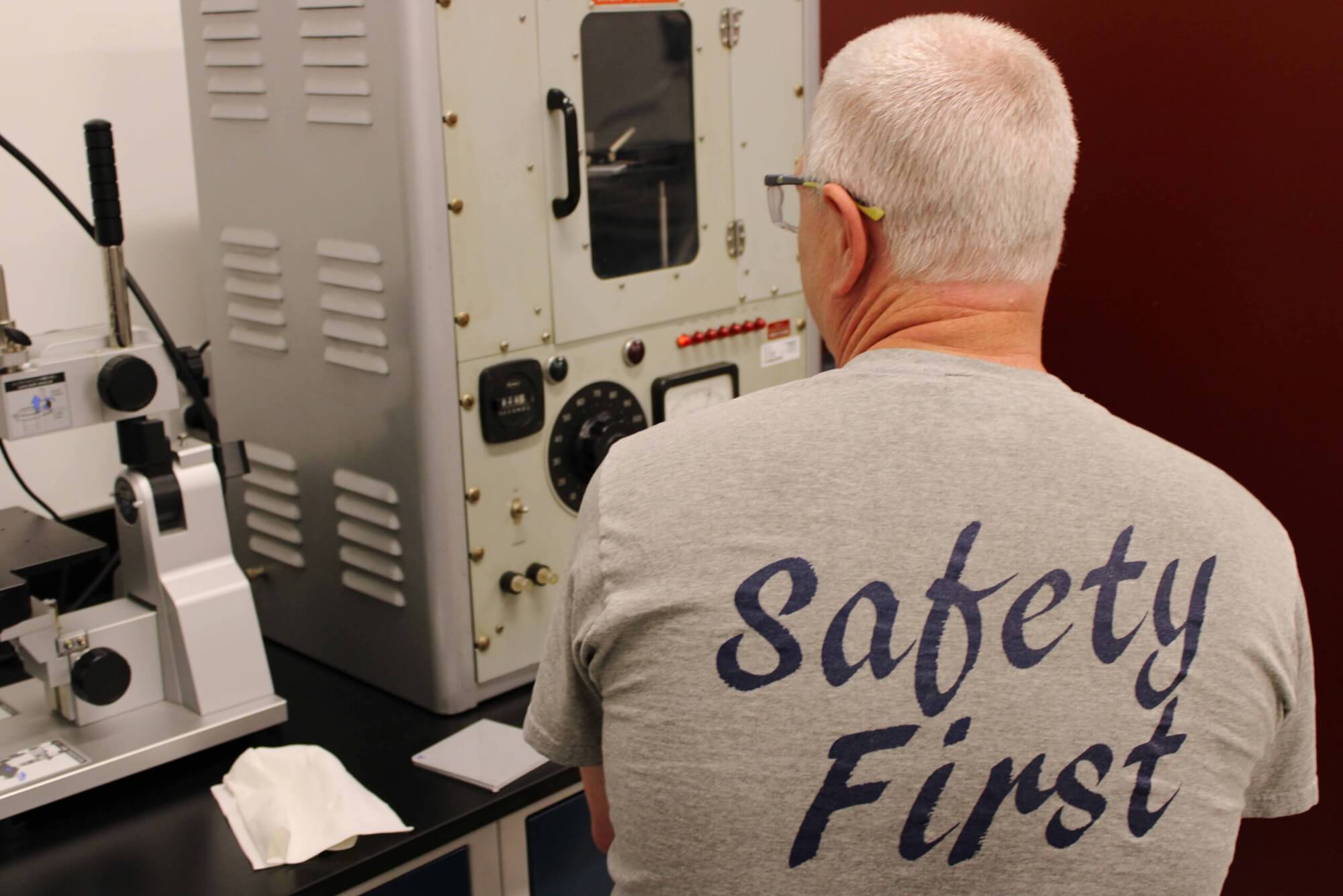 A person sitting in front of a lab machine with a shirt on that has "safety first" text on the back of the shirt and safety glasses on.
