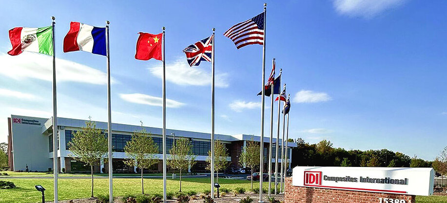 A picture of IDI Composites International outside with the flags blowing in the forefront and the large manufacturing facility in the background and blue skies.