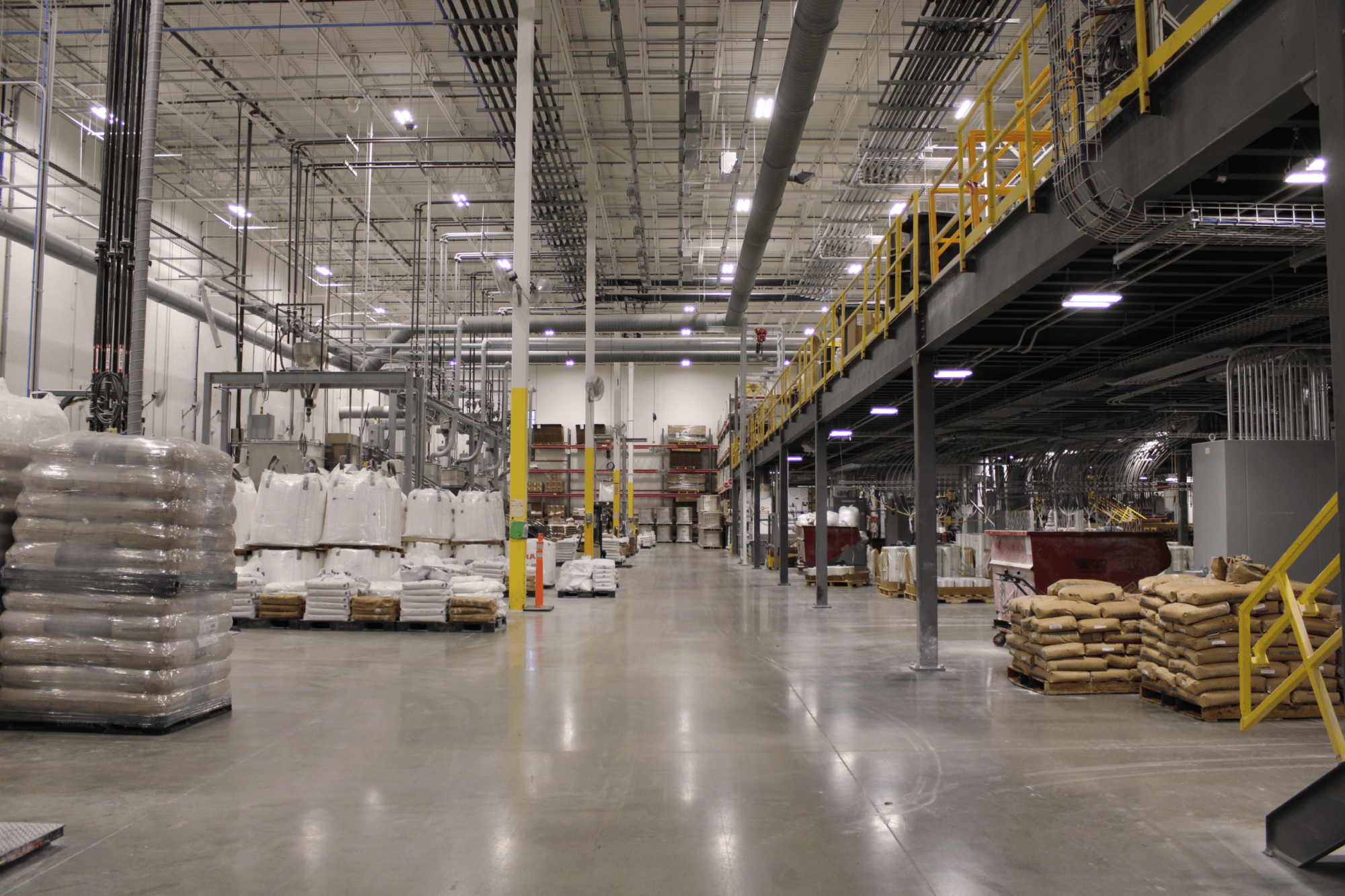 A manufacturing floor with bags of raw materials on the right side and a grey and yellow mezzanine on the left side with other raw materials underneath the mezzanine.