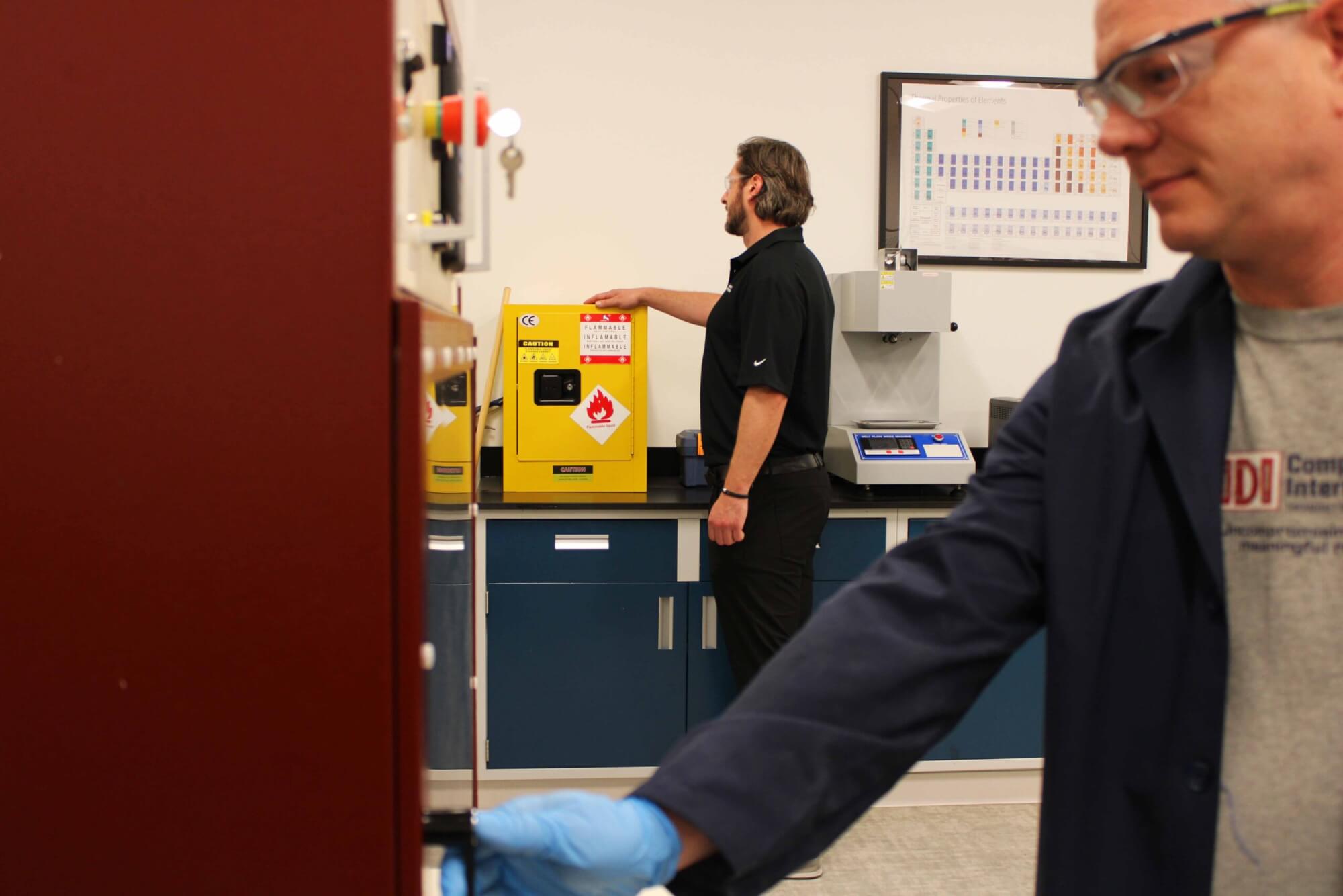 Two people in a lab environment. One person is wearing a shirt that says IDI Composites International and a longer navy lab coat and the other peson is wearing all black and touching a yellow lab machine.